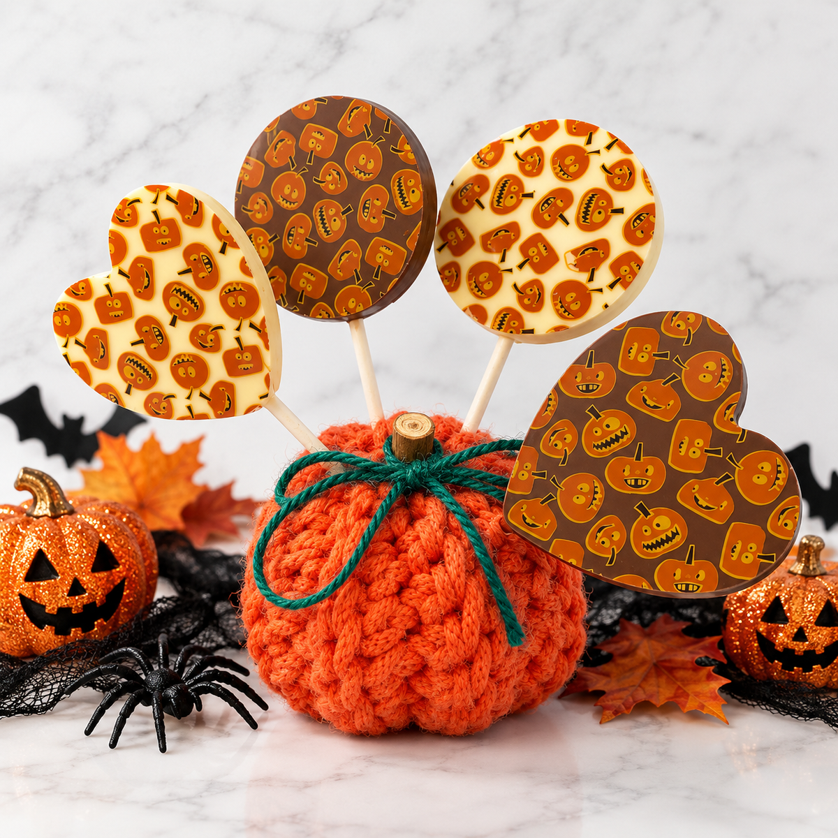 Halloween-themed lollipop stand featuring a crocheted orange pumpkin base with a green bow, holding four patterned lollipops. The stand is styled with miniature jack-o'-lanterns, black spiderwebs, black plastic spiders, and orange autumn leaves on a white marble surface.